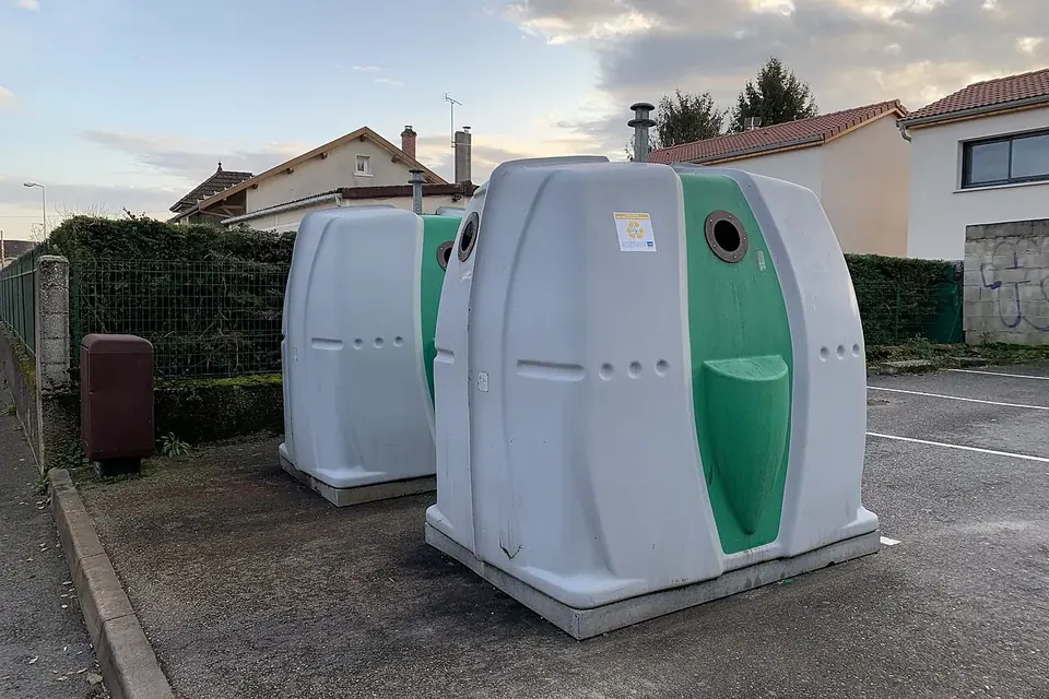 Deux conteneurs à verre en plastique blanc et vert posés sur un parking, devant des maisons